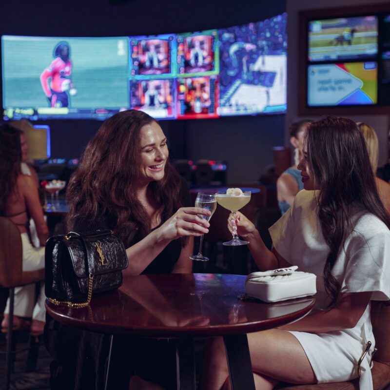 Two women enjoying cocktails at Sports Bar inside the casino, with screens and a lively atmosphere